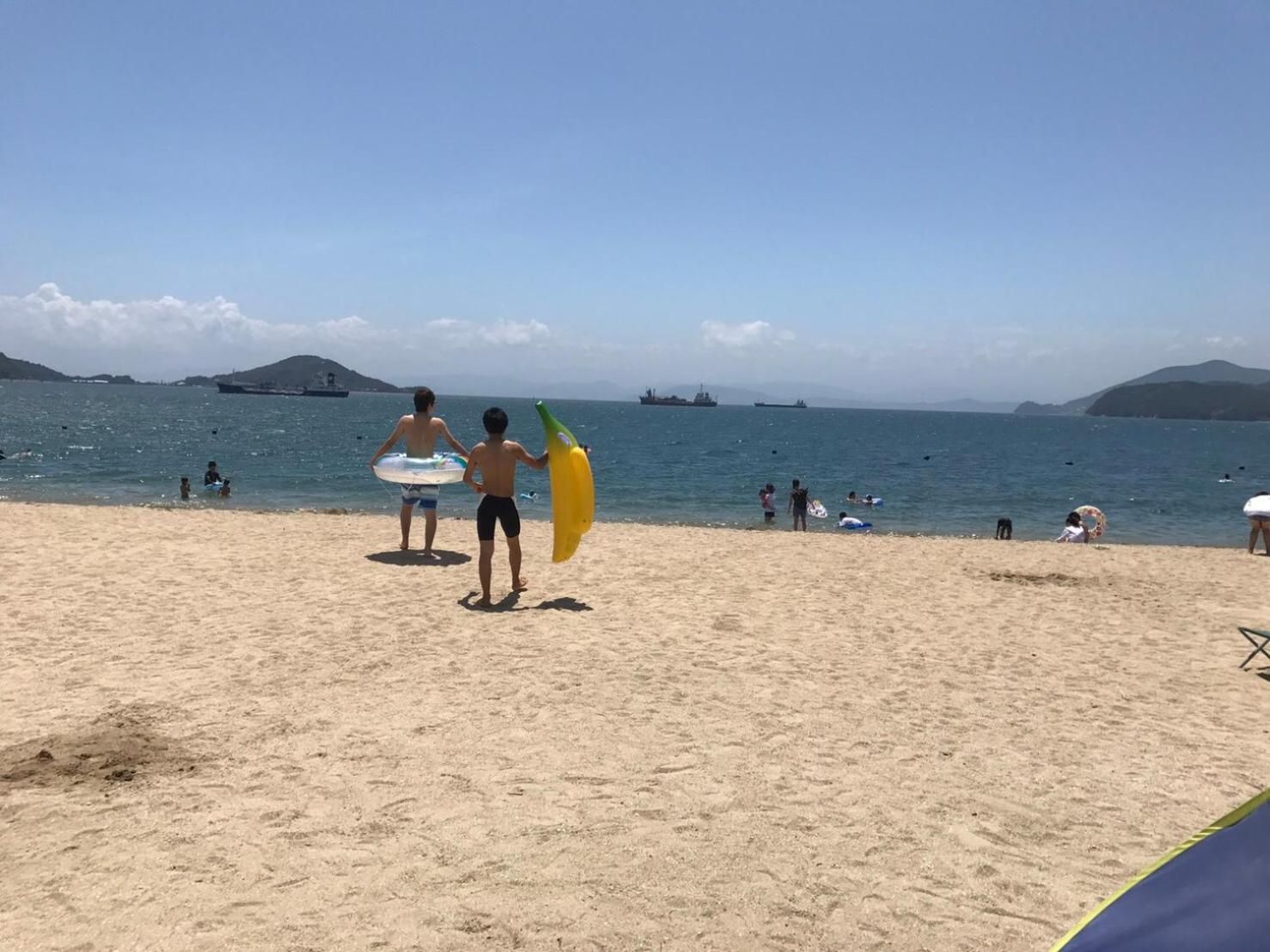 A beach with people, some holding surfboards, and ships in the distance.