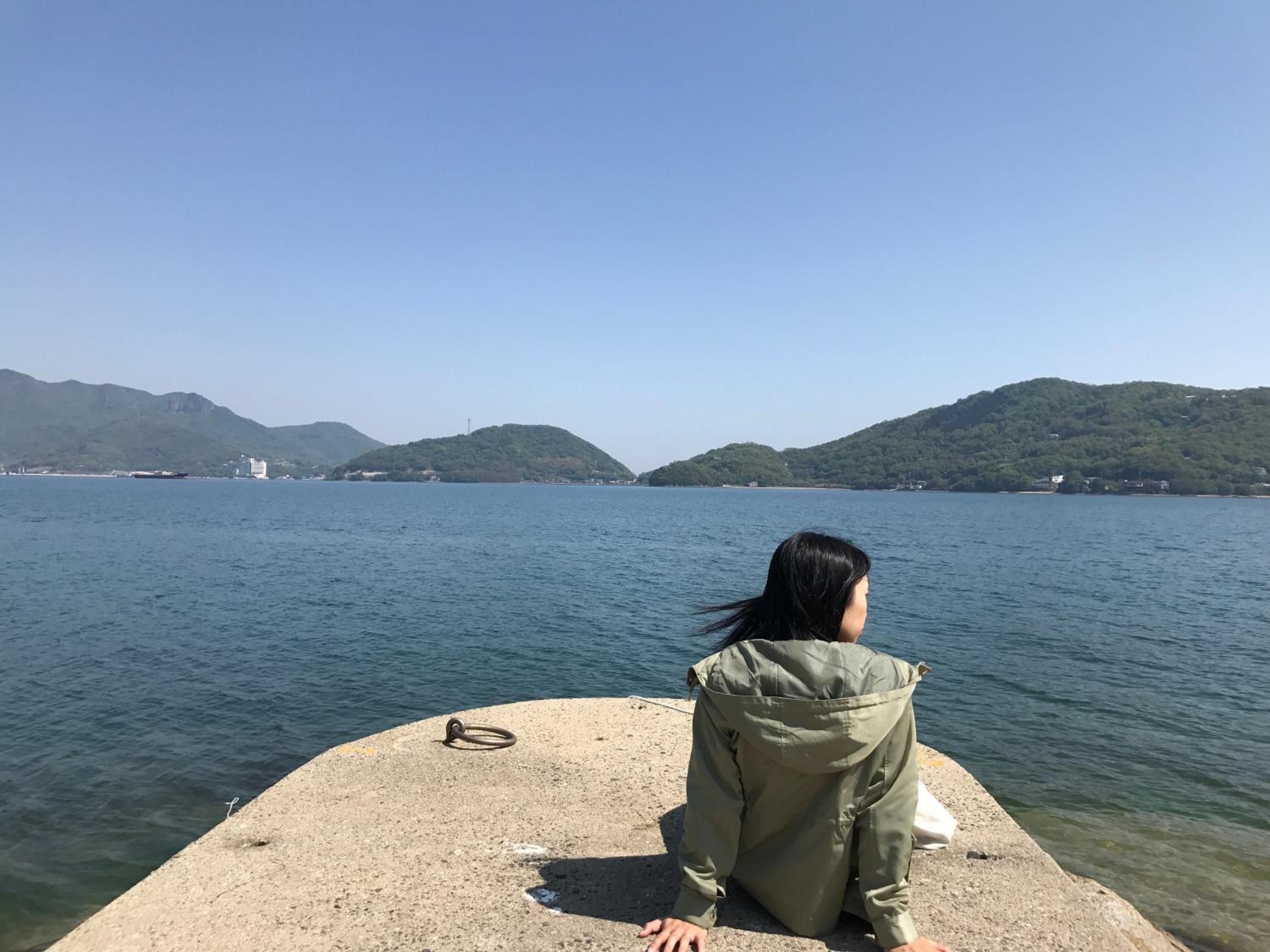 A person sitting on a concrete structure by a body of water with hills in the background.