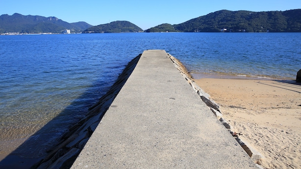 Un quai en béton s’étendant dans une étendue d’eau calme, avec un ciel dégagé et des montagnes en arrière-plan.