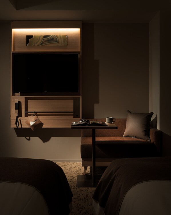 Habitación de hotel tenuemente iluminada, con un televisor de pantalla plana en la pared, una mesita con una taza y un libro, y un sofá con un cojín.