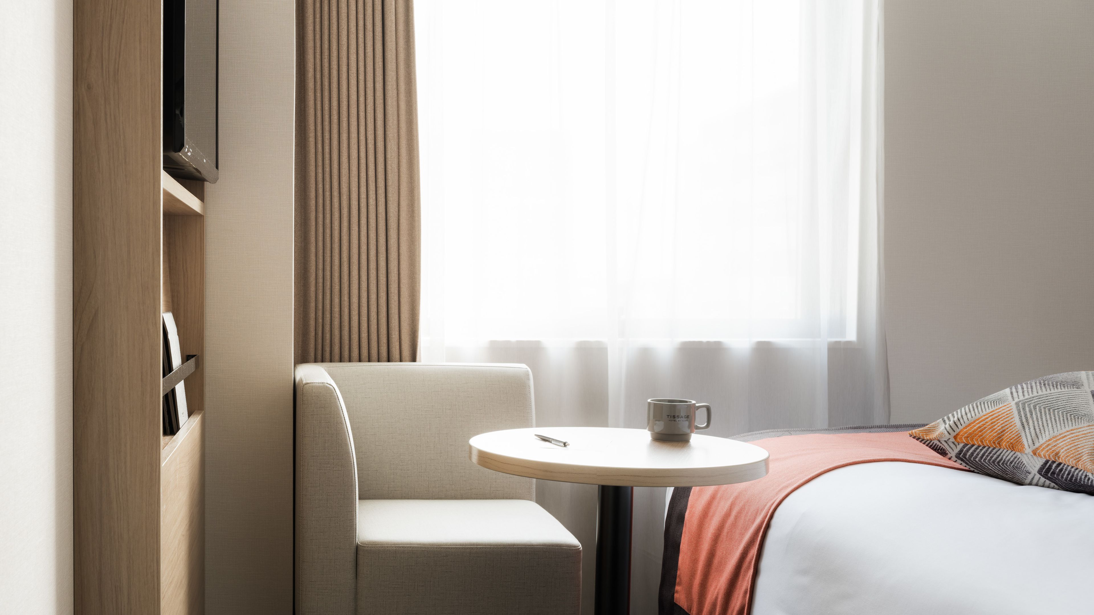A hotel room with a single bed, a small round table with a cup, a chair, and a window with sheer curtains.