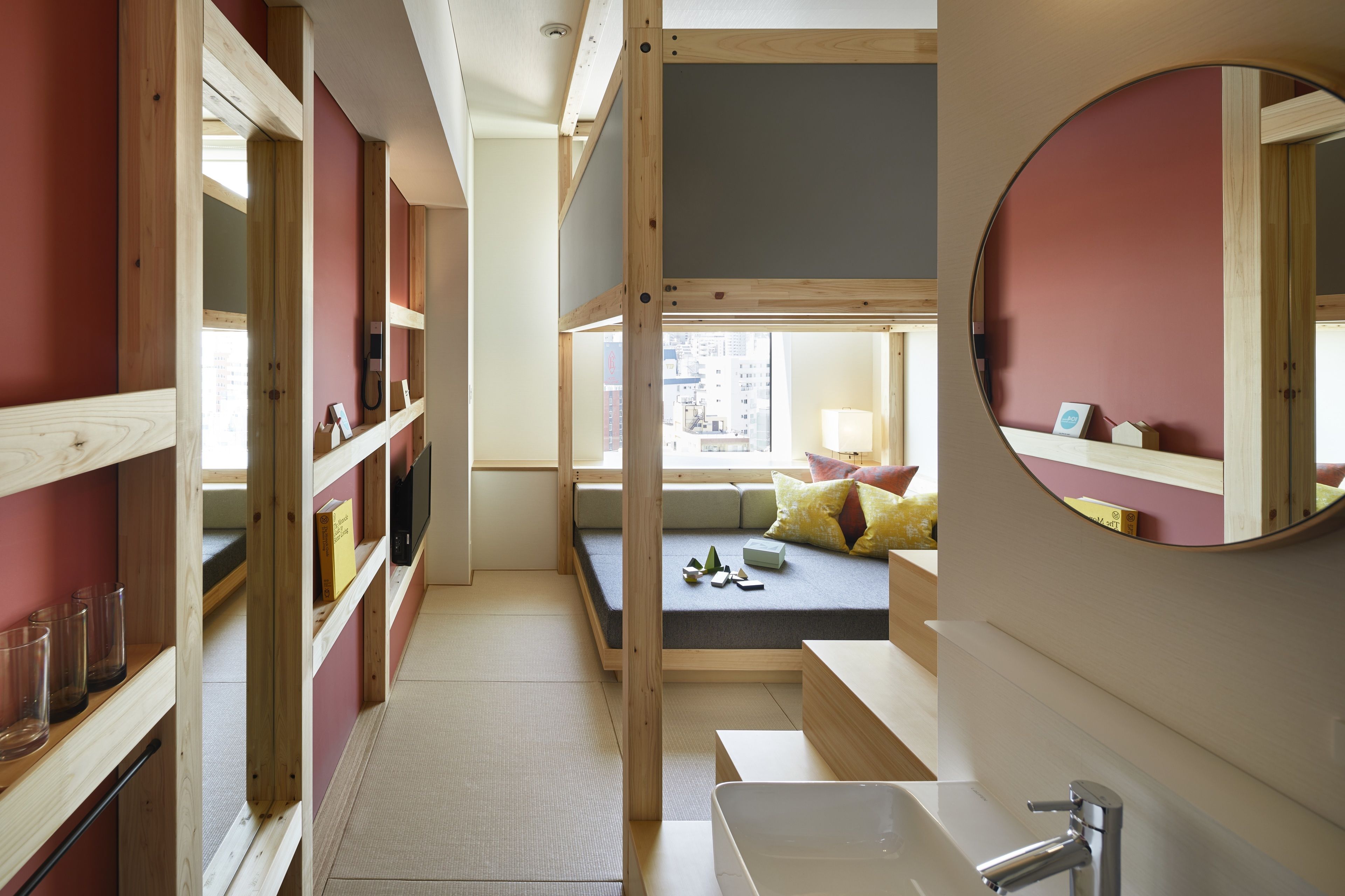 Une chambre d’hôtel compacte avec un lit intégré, un petit coin salon, une étagère en bois avec des verres et un miroir rond.