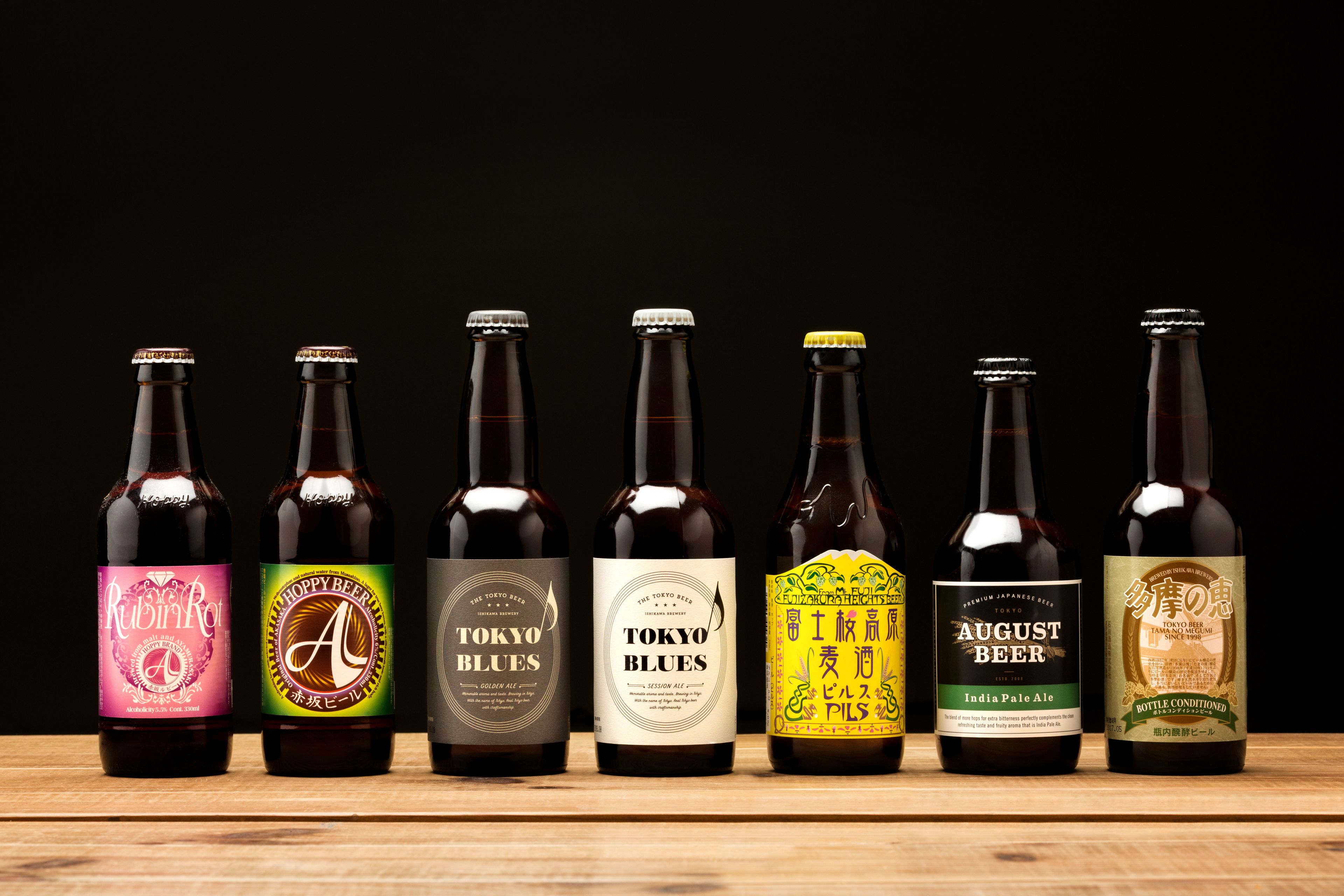 Sept bouteilles de bière, toutes étiquetées, disposées en rangée sur une surface en bois, le tout sur un fond sombre.