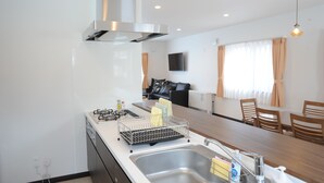 Una cocina moderna con extractor de acero inoxidable, lavavajillas y fregadero. Hay una zona de comedor con sillas de madera y una mesa, y una sala de estar con un sofá negro y un televisor montado en la pared.