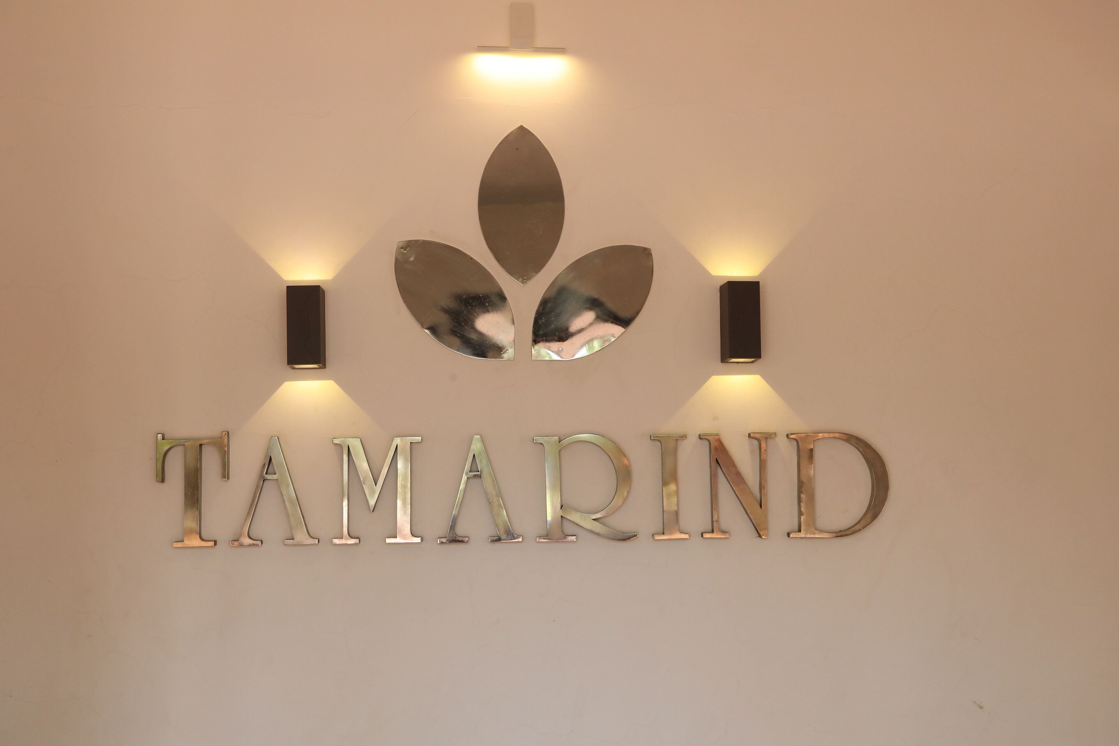 A wall-mounted sign with the name "TAMARIND" in metallic letters, flanked by two wall-mounted lights and a decorative leaf-shaped mirror above.