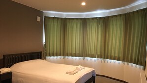 A hotel room with a bed, a nightstand, a lamp, and a window with green curtains. - Jadan House (Furano)