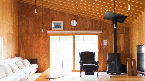 Ein gemütlicher Raum mit Holzboden, einem schwarzen Stuhl, einem Tisch, einer Couch und einem Kaminofen.