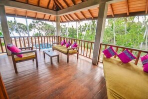 Una terraza de madera con una mesa de madera y dos sillas, un banco acolchado con cojines y vistas a una piscina y vegetación tropical.