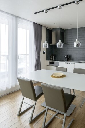A modern kitchen with a white table and chairs, a coffee machine, and a view of the outdoors through large windows.