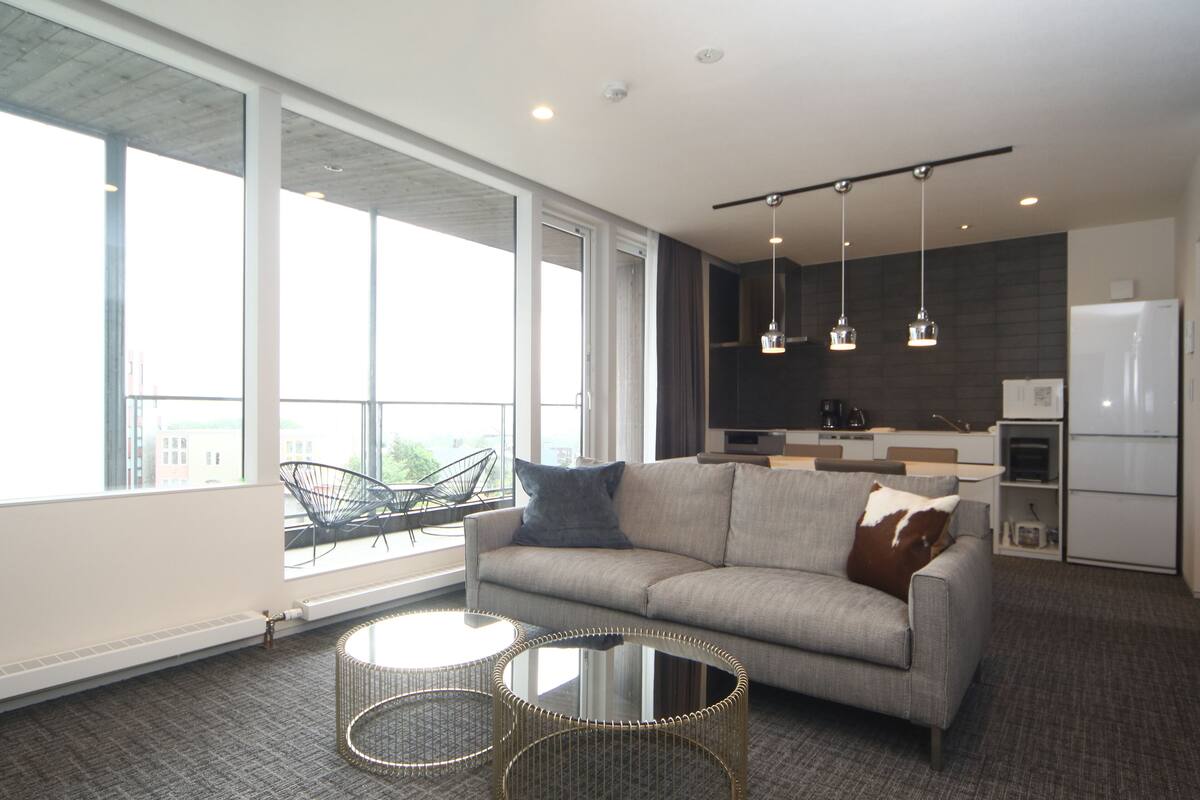 A modern living room with a grey sofa, two round glass tables, and a large window offering a city view.