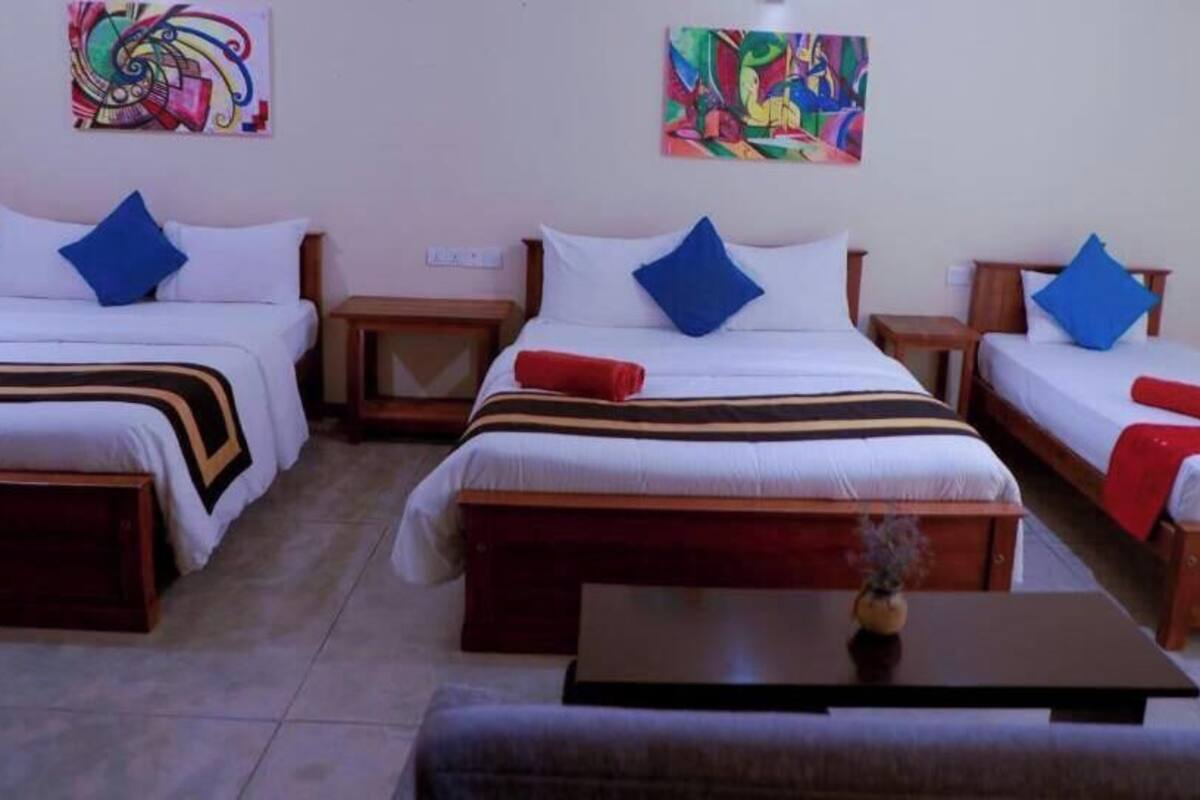 A hotel room with three single beds, each with a wooden frame and white bedding. There are colorful abstract paintings on the walls, a small table with a vase, and a bench at the foot of the beds.