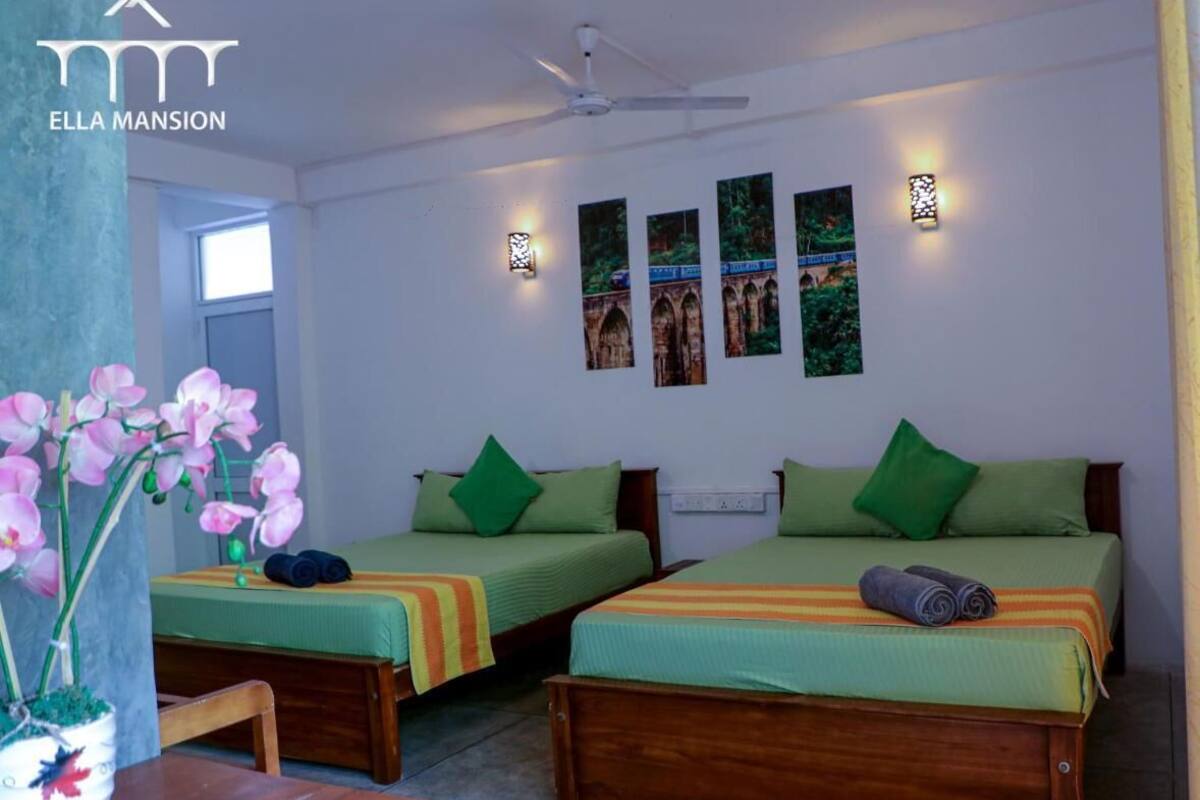 A hotel room with two single beds, green bedding, a small table with a chair, a potted orchid, and wall-mounted artwork.