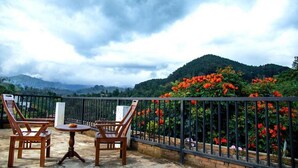 A balcony with two wooden chairs and a small table, overlooking a lush garden with vibrant red flowers and a mountainous landscape under a cloudy sky. - Ella Mansion (Ella)