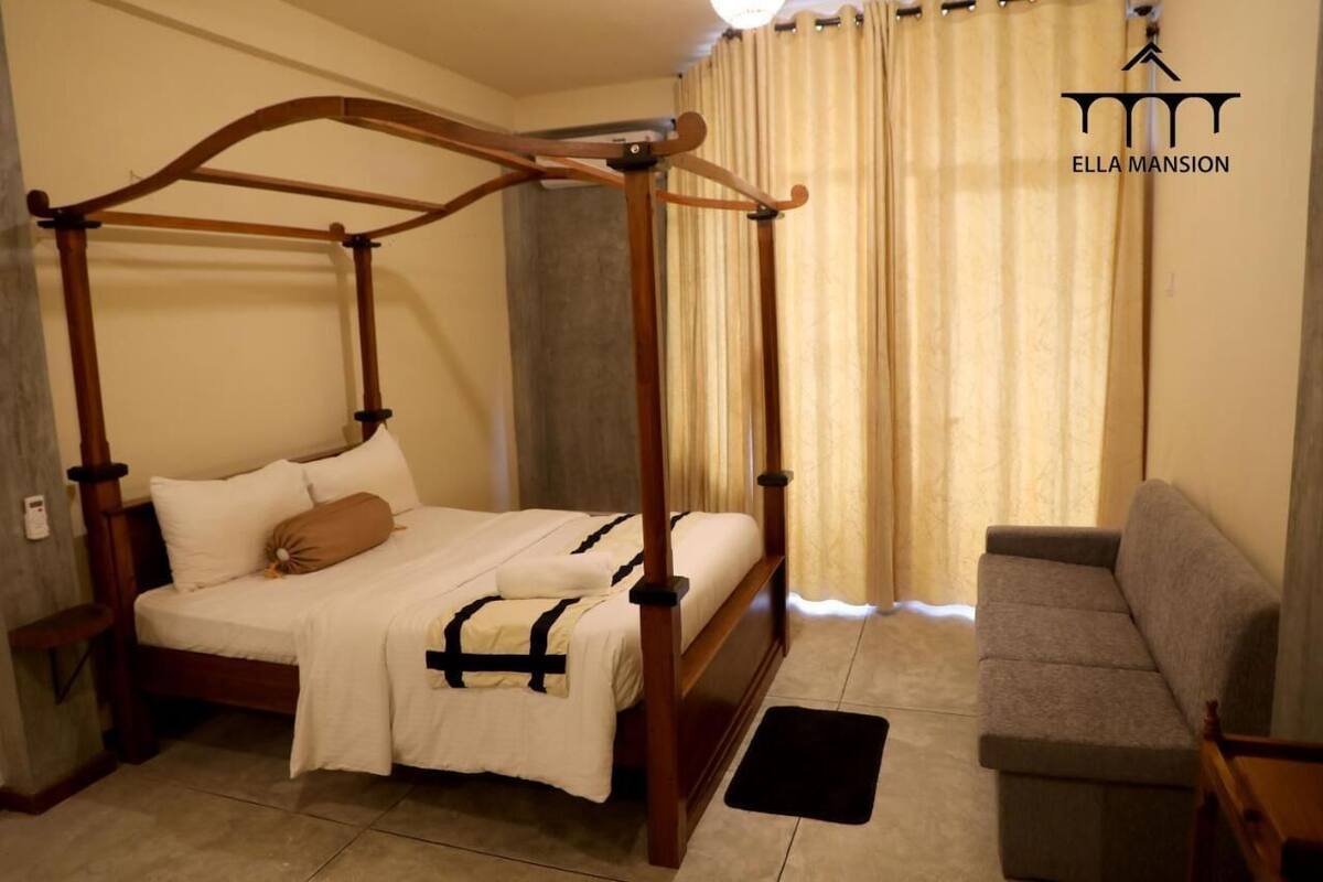 A four-poster bed with white bedding and a brown pillow, a grey sofa, and a black rug in a room with beige curtains and a light fixture.