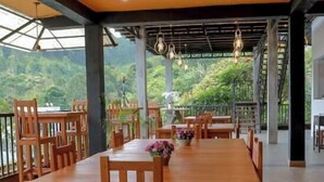 A spacious dining area with wooden tables and chairs, large windows offering a view of lush greenery, and hanging pendant lights. - Ella Mansion (Ella)