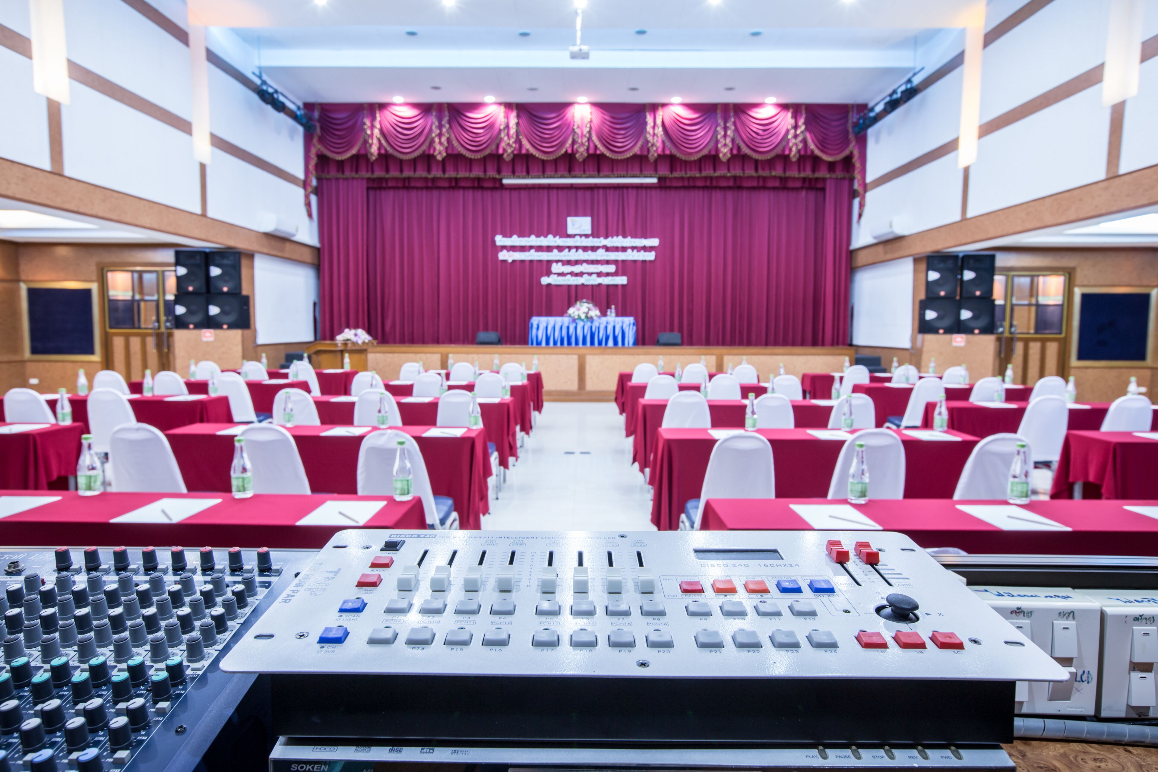 A conference room with rows of tables and chairs, a stage with a red curtain, and a sound mixing console in the foreground.