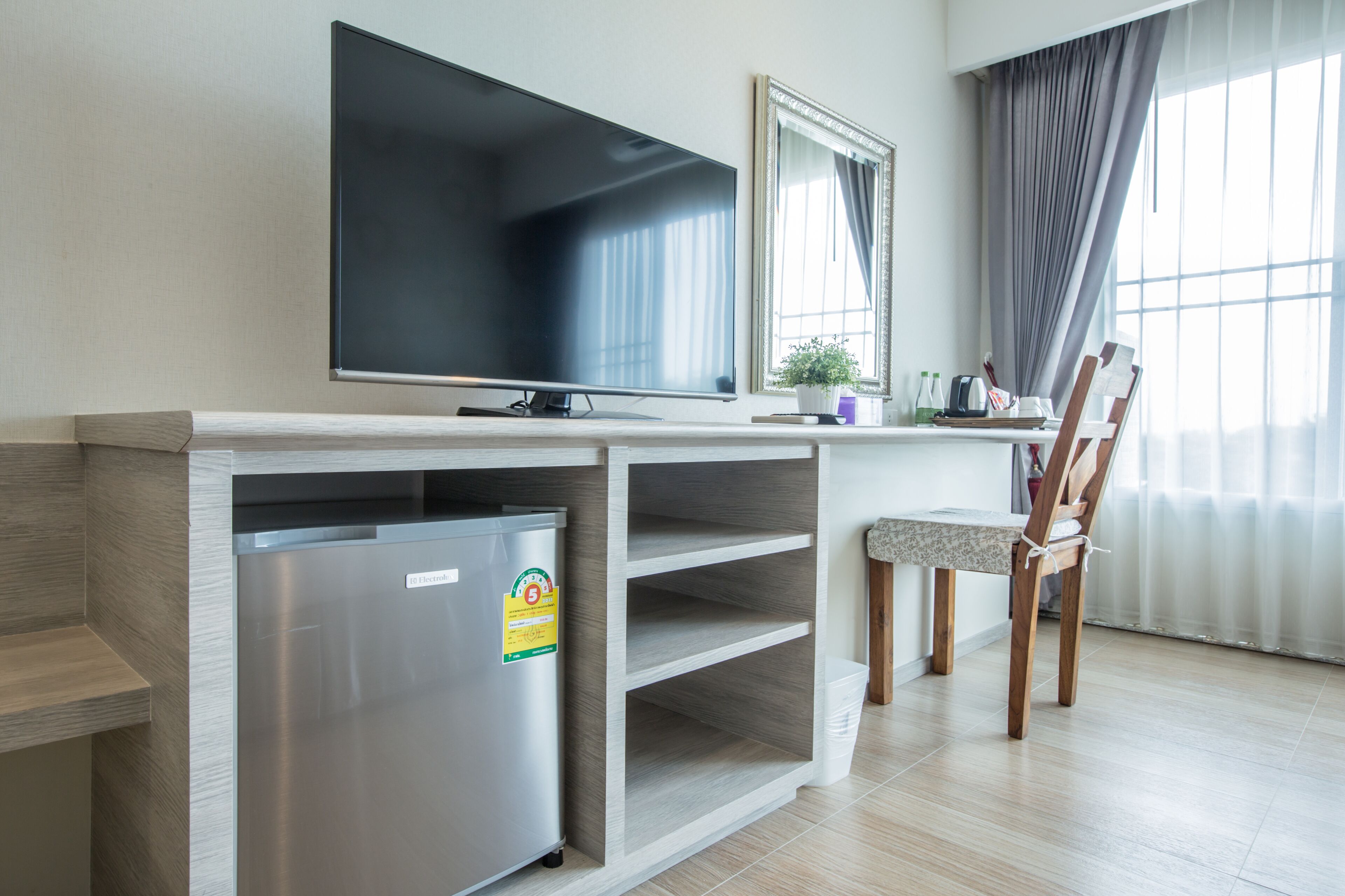 A modern hotel room with a flat-screen TV, a mini-fridge, a wooden desk, a chair, and a large window with curtains.