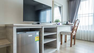 A modern hotel room with a flat-screen TV, a mini-fridge, a wooden desk, a chair, and a large window with curtains.