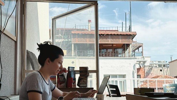 Una persona trabajando en una mesa con una computadora portátil, una taza de café y una ventana que da a vistas de edificios.