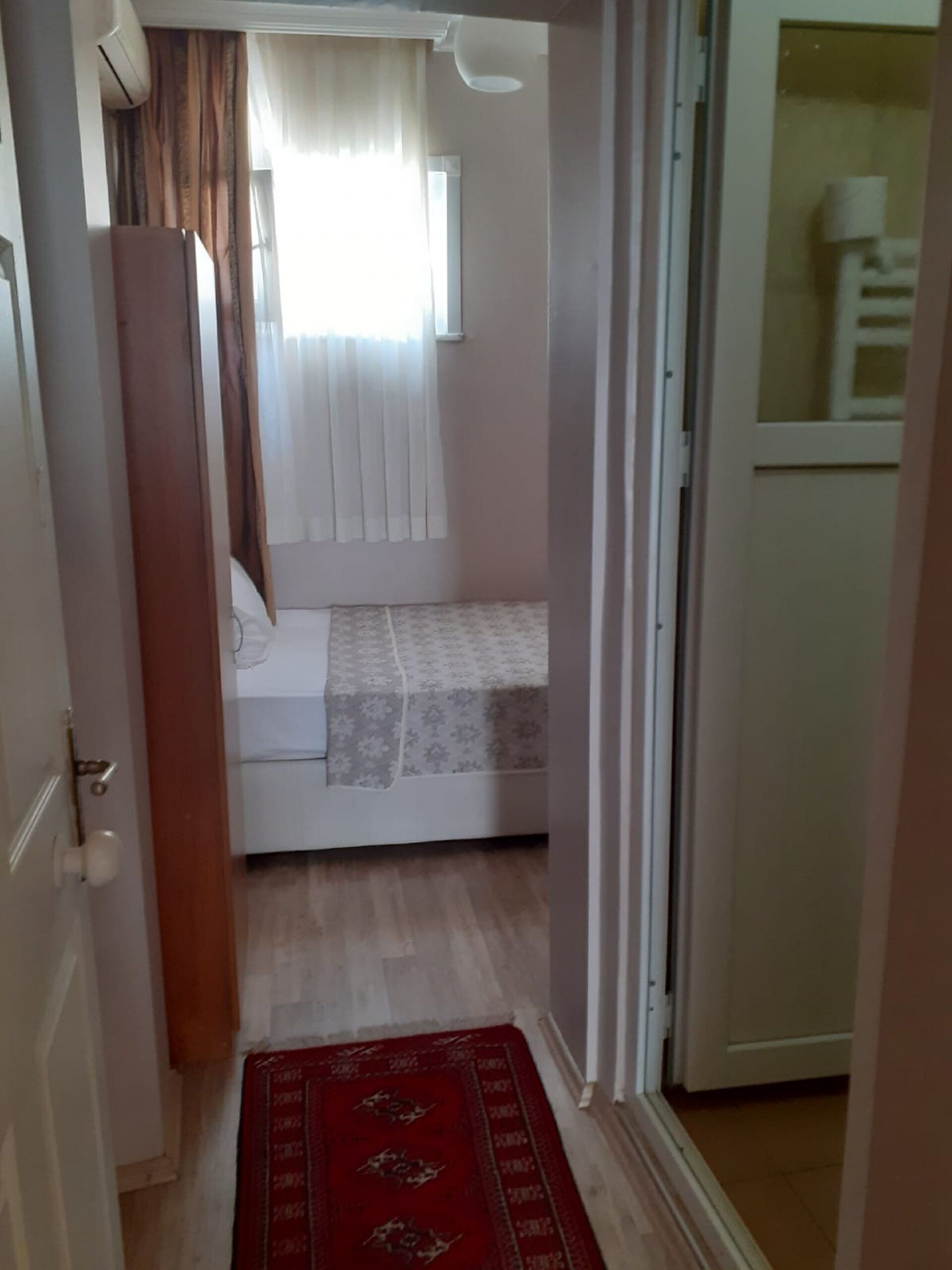 Een smalle gang die leidt naar een slaapkamer met een bed, een raam met gordijnen en een rode vloerkleed.