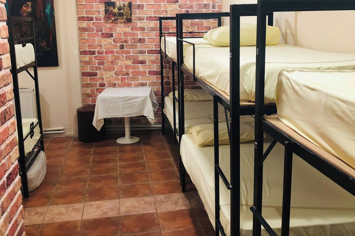A dormitory room with bunk beds, a brick wall, a small table with a white tablecloth, and a painting on the wall.