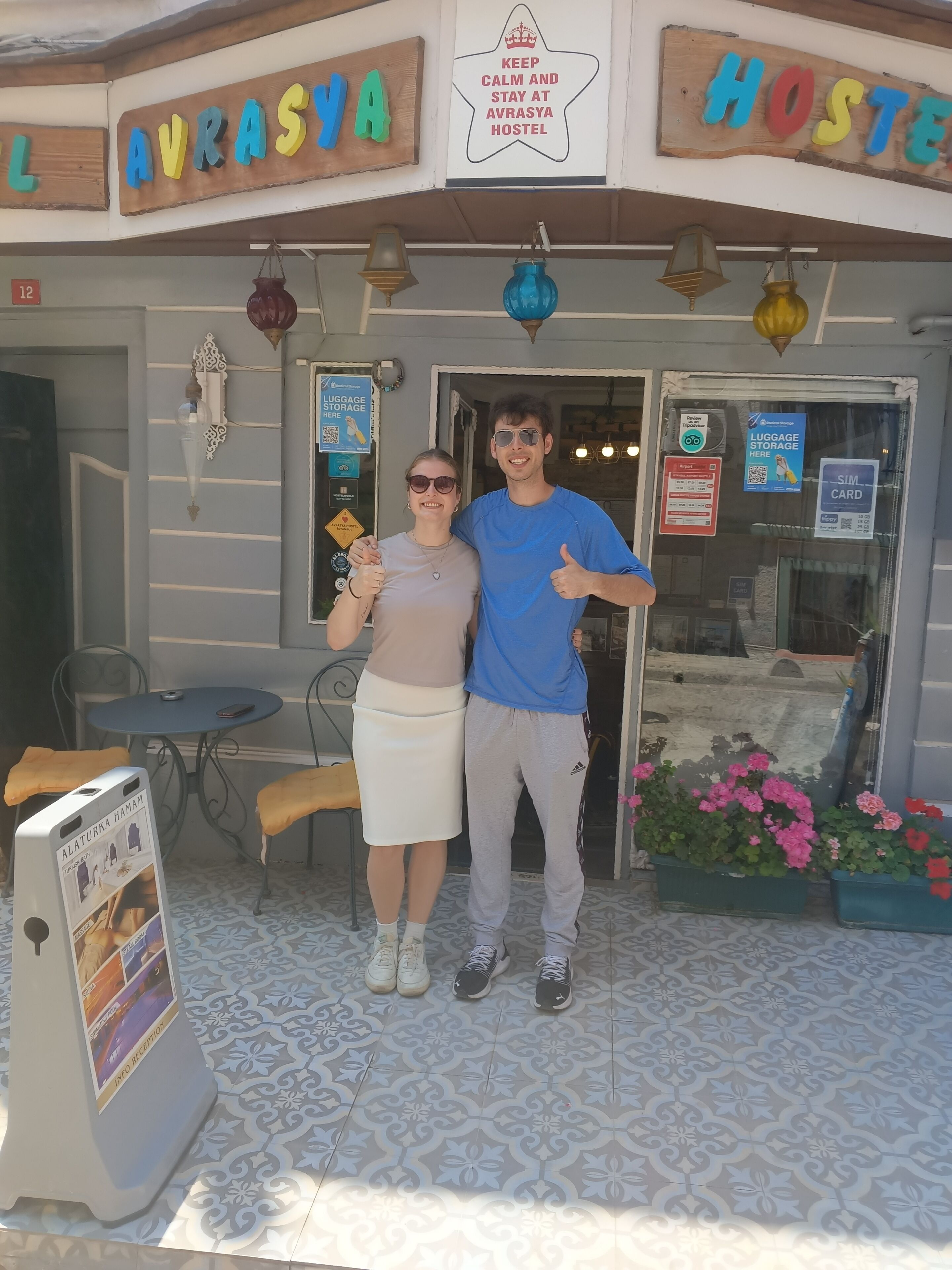 A hostel with a sign reading 'AVRASYA HOSTEL' and 'KEEP CALM AND STAY AT AVRASYA HOSTEL.' There is a luggage storage sign, a SIM card sign, and a small table with a chair outside.