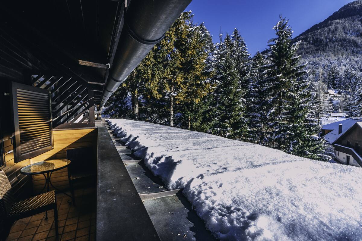 Un espacio al aire libre cubierto de nieve con un banco y sillas de madera, una barandilla metálica y árboles nevados al fondo.