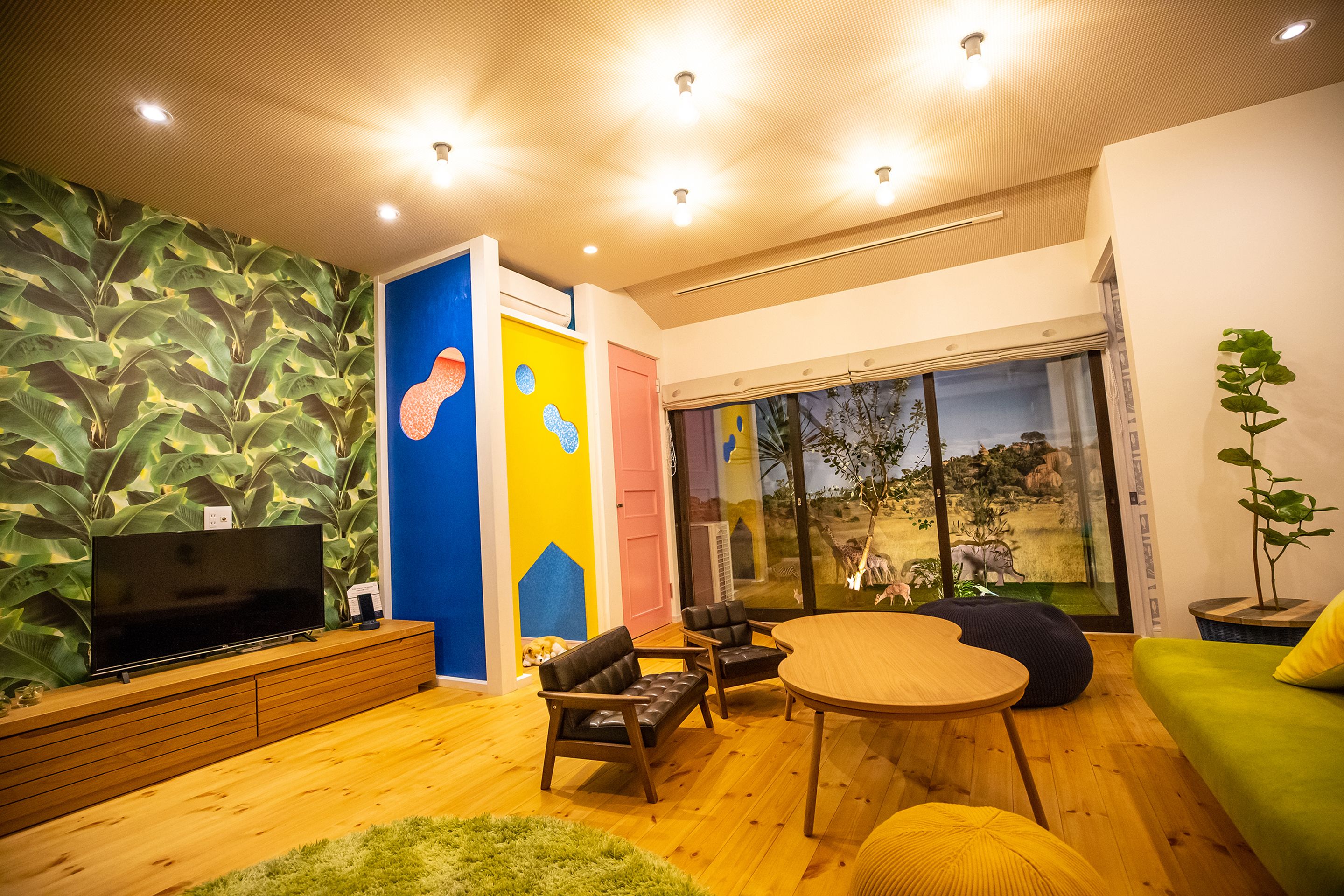 A modern living room with a flat-screen TV, wooden furniture, a large window with a view of a park, and a vibrant wall mural.