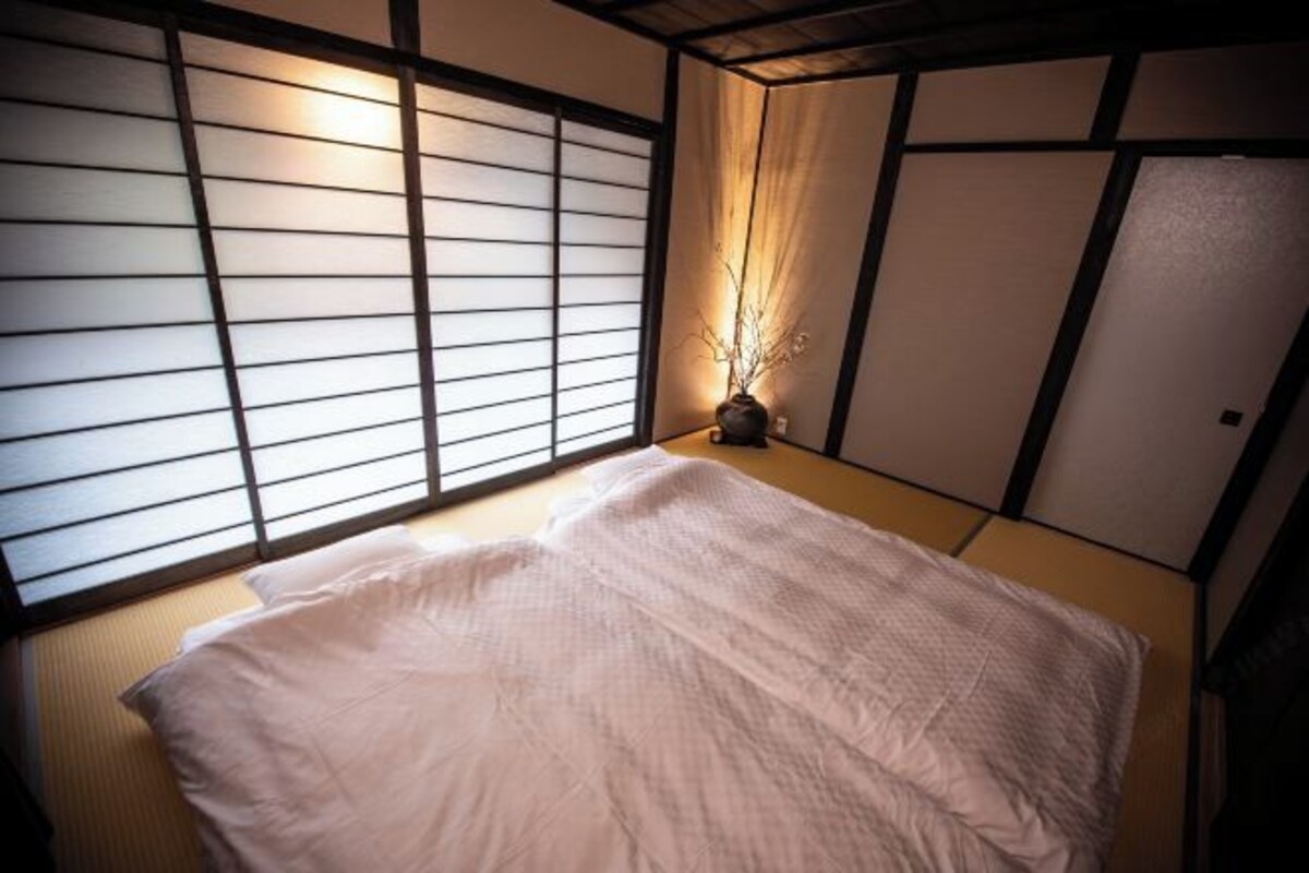Een traditionele Japanse kamer met tatami vloerbedekking, een laag bed, shoji schermen en een kleine vaas met takken.