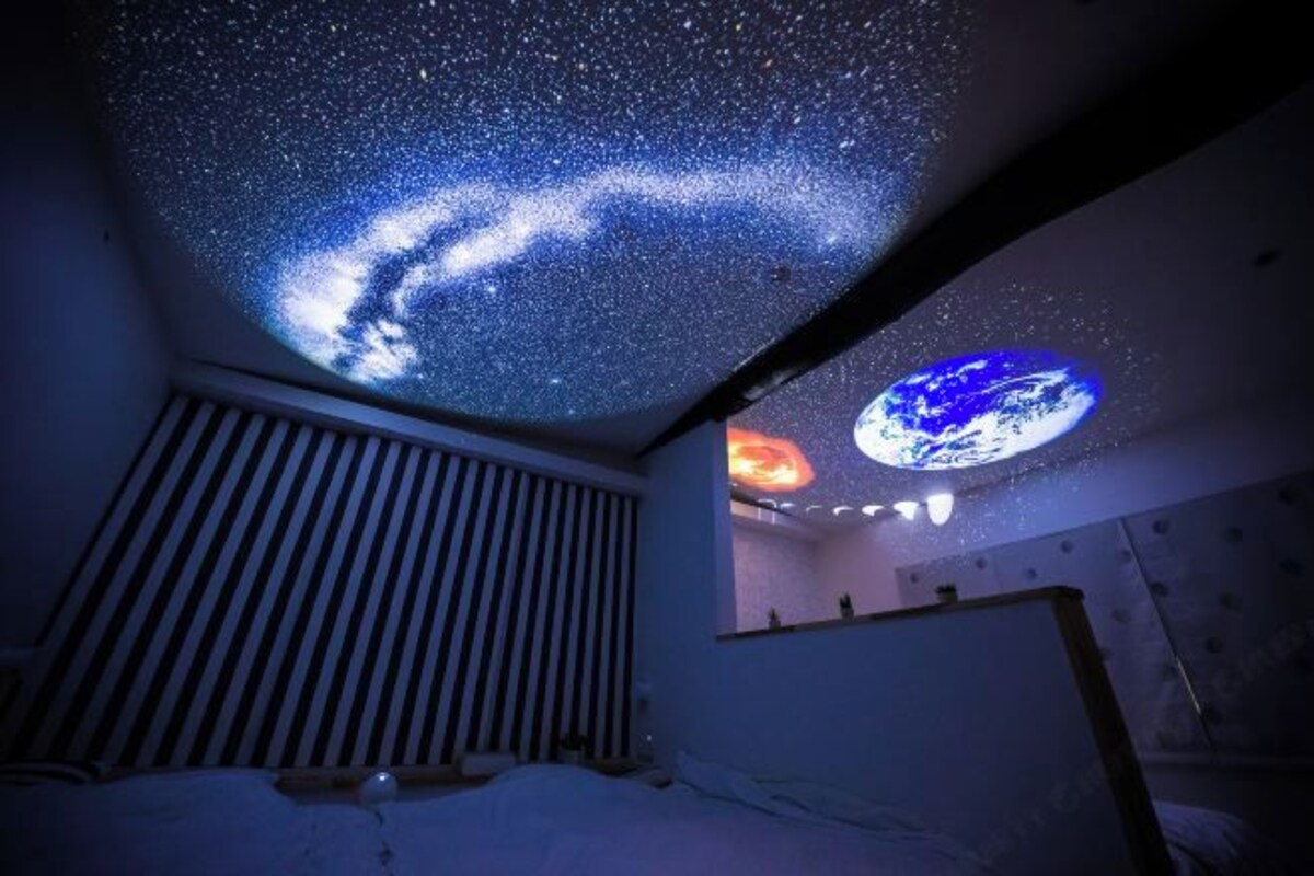 Een kamer met een projectie van een sterrenhemel op het plafond, een bed en een raam met uitzicht op de aarde.