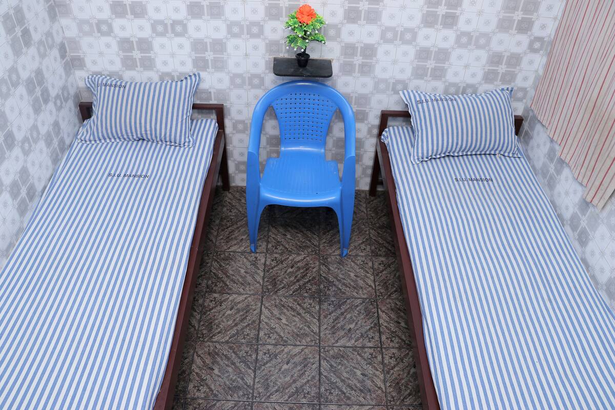 Una habitación con dos camas individuales, cada una con ropa de cama a rayas, una silla de plástico azul y una mesita con una planta en maceta.