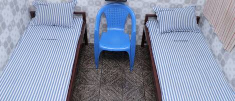 A room with two single beds, each with striped bedding, a blue plastic chair, and a small table with a potted plant.