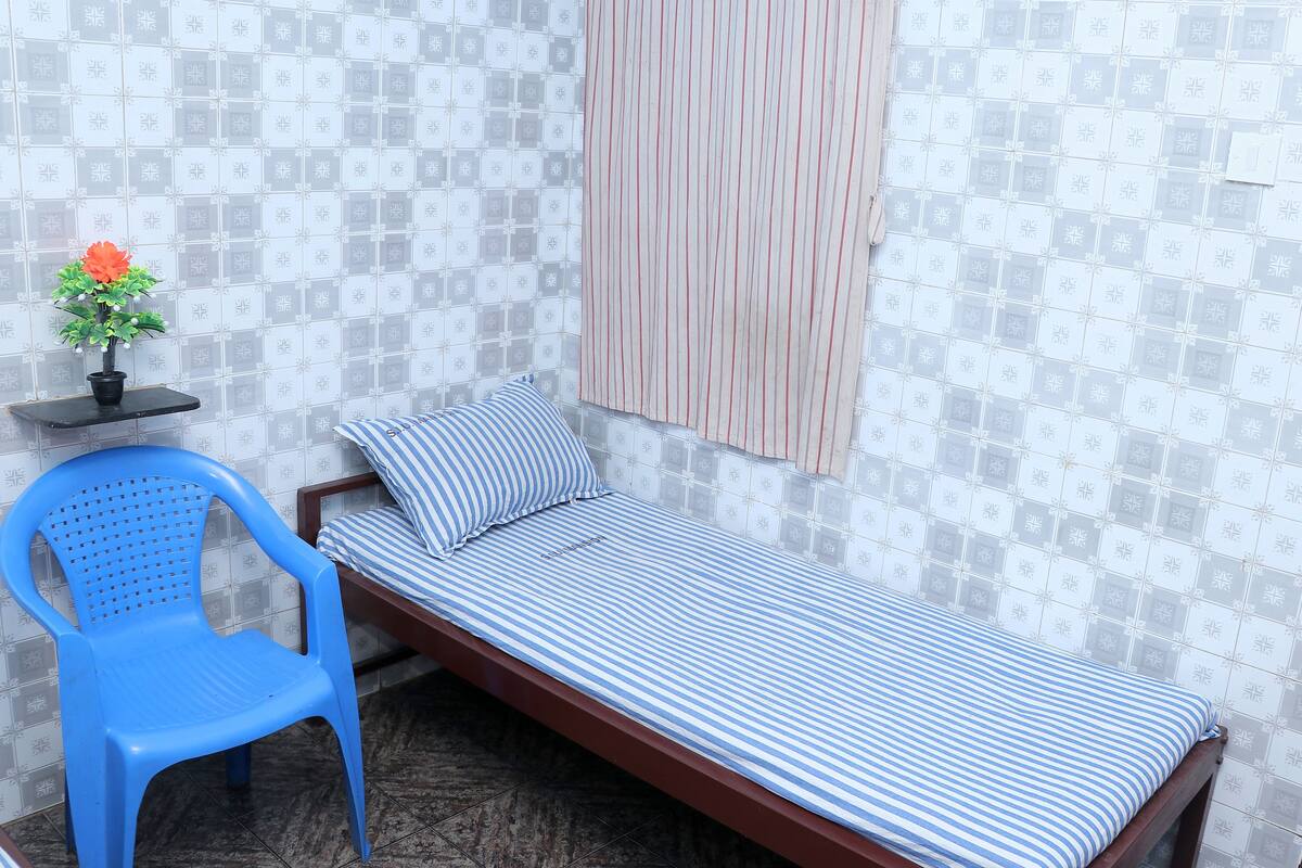 Una habitación pequeña con una cama individual, una colcha a rayas, una silla de plástico azul, una mesita con una planta en maceta y una cortina a rayas.