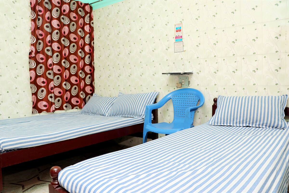 Una habitación con dos camas individuales, una silla de plástico azul y cortinas con estampado.