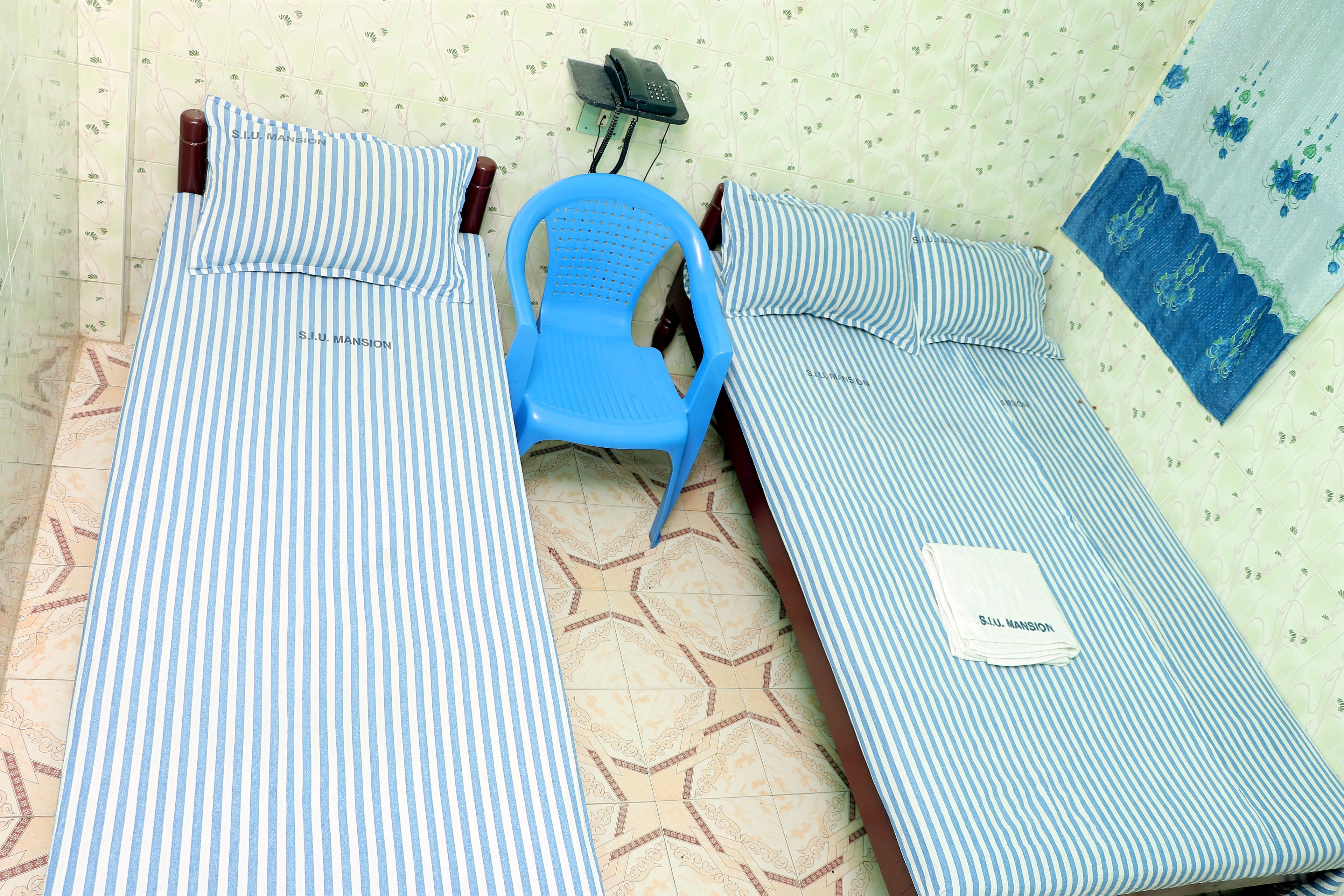 Dos camas individuales con ropa de cama a rayas, una silla de plástico azul y un teléfono en la pared.