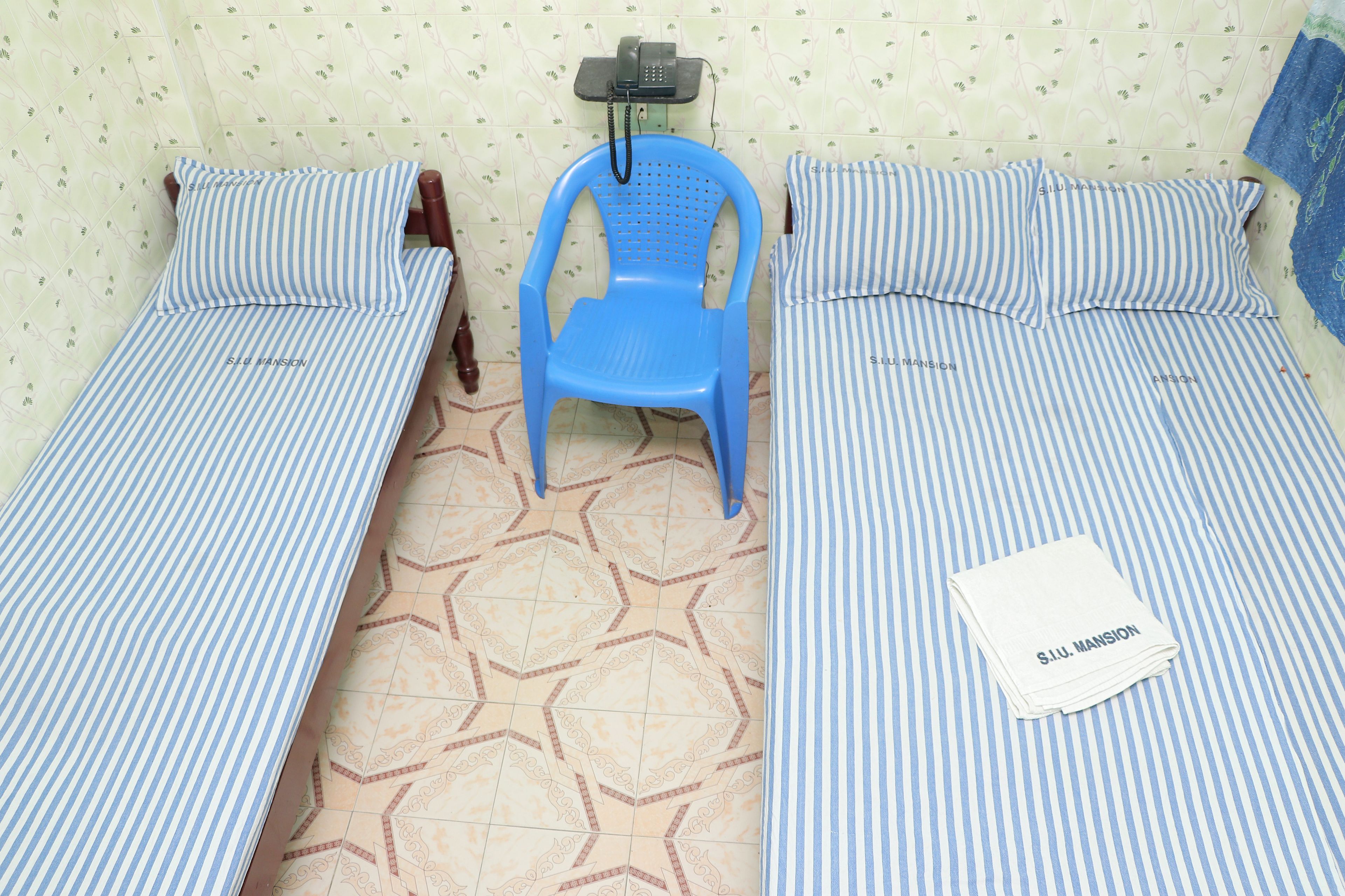 Two single beds with striped bedding, a blue plastic chair, a telephone on a stand, and a folded white towel on one bed.