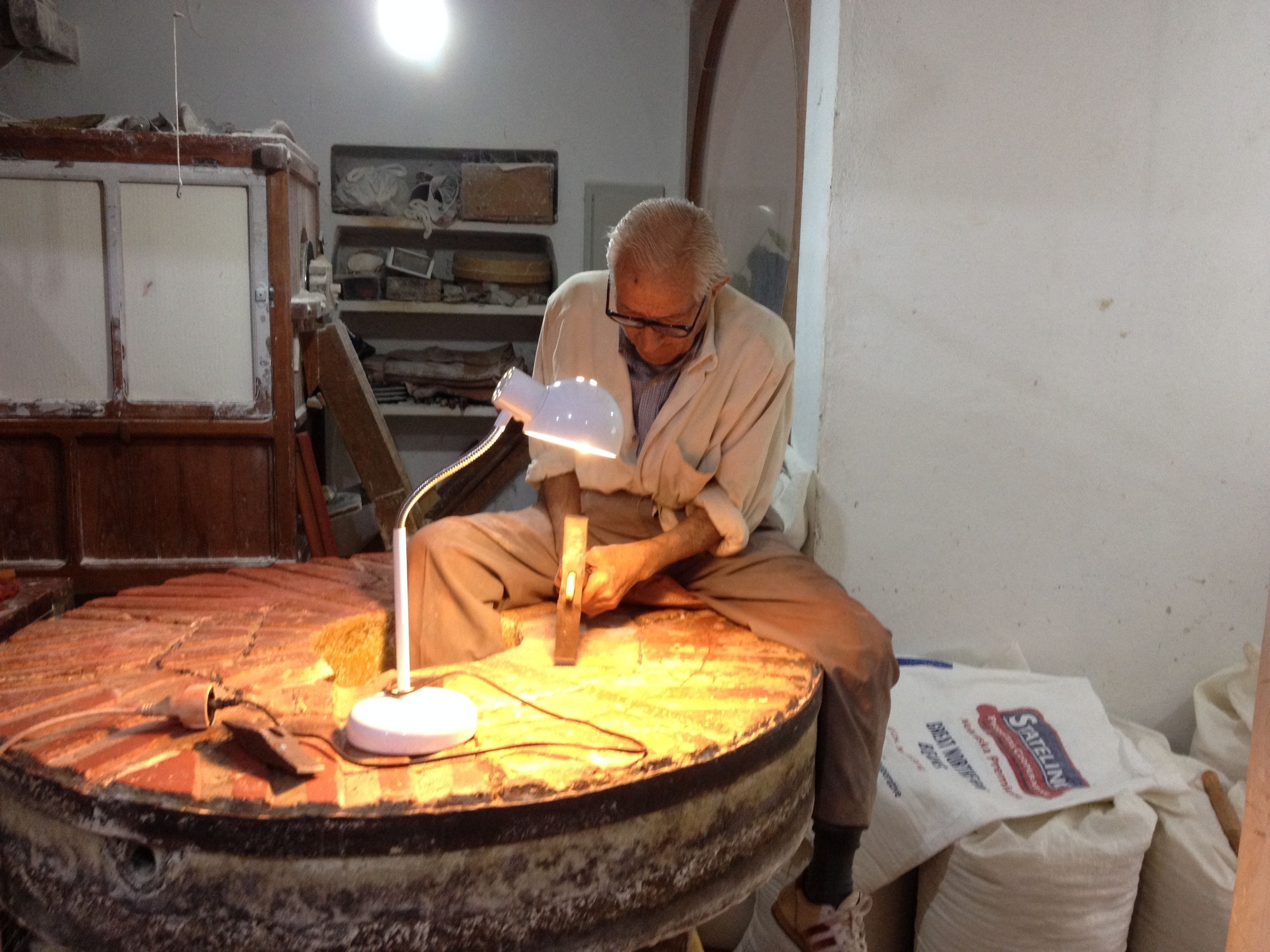 Una persona trabajando en una mesa de madera con una lámpara, rodeada de diferentes objetos y un saco de harina.