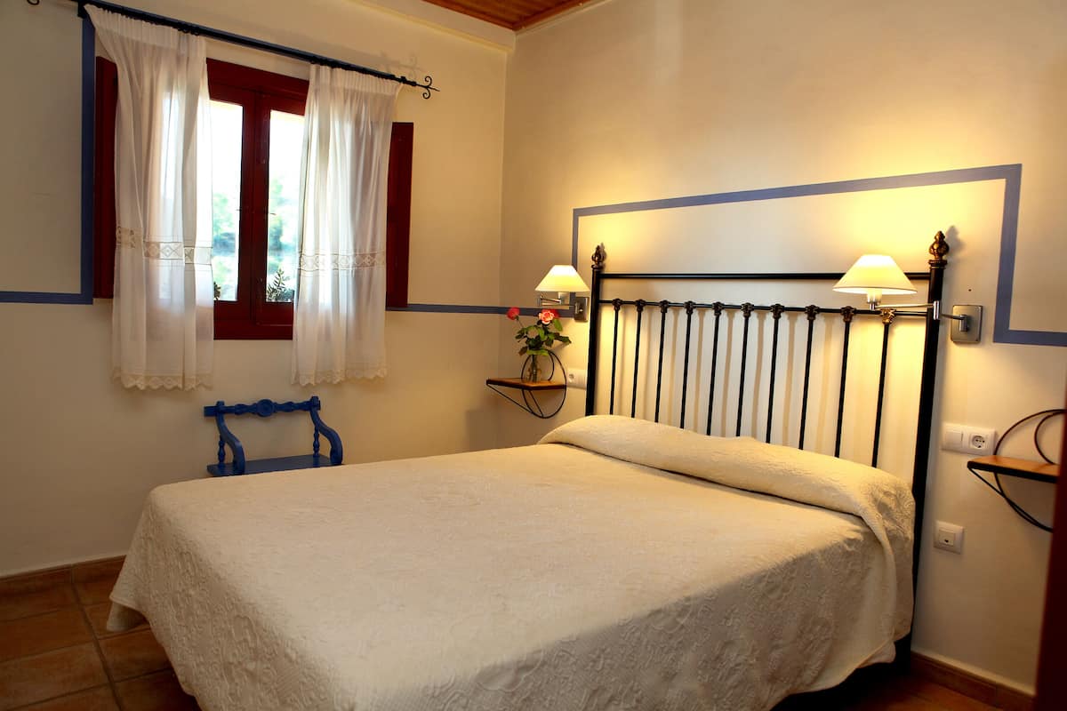 Una cama bien hecha con una colcha blanca, una mesita de noche con una lámpara y una ventana con cortinas.