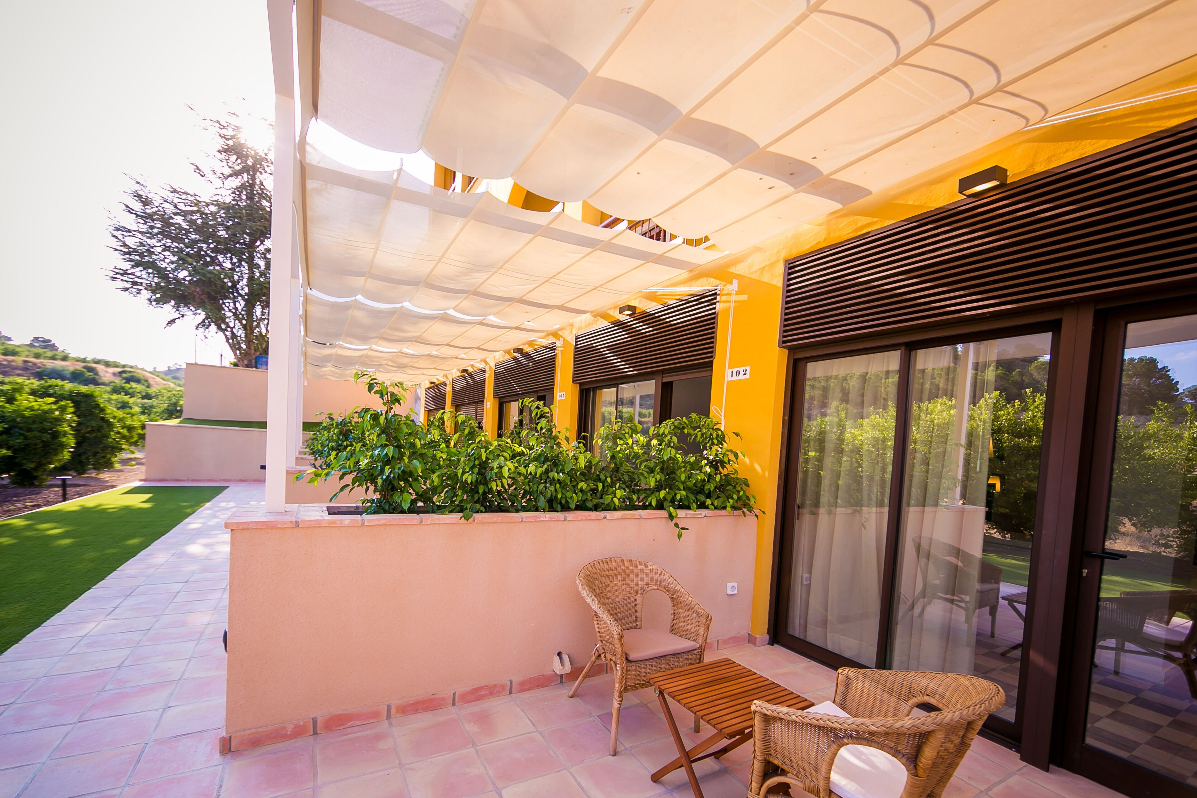 A covered outdoor seating area with wicker furniture, a glass door leading to a patio, and a view of the surrounding greenery.