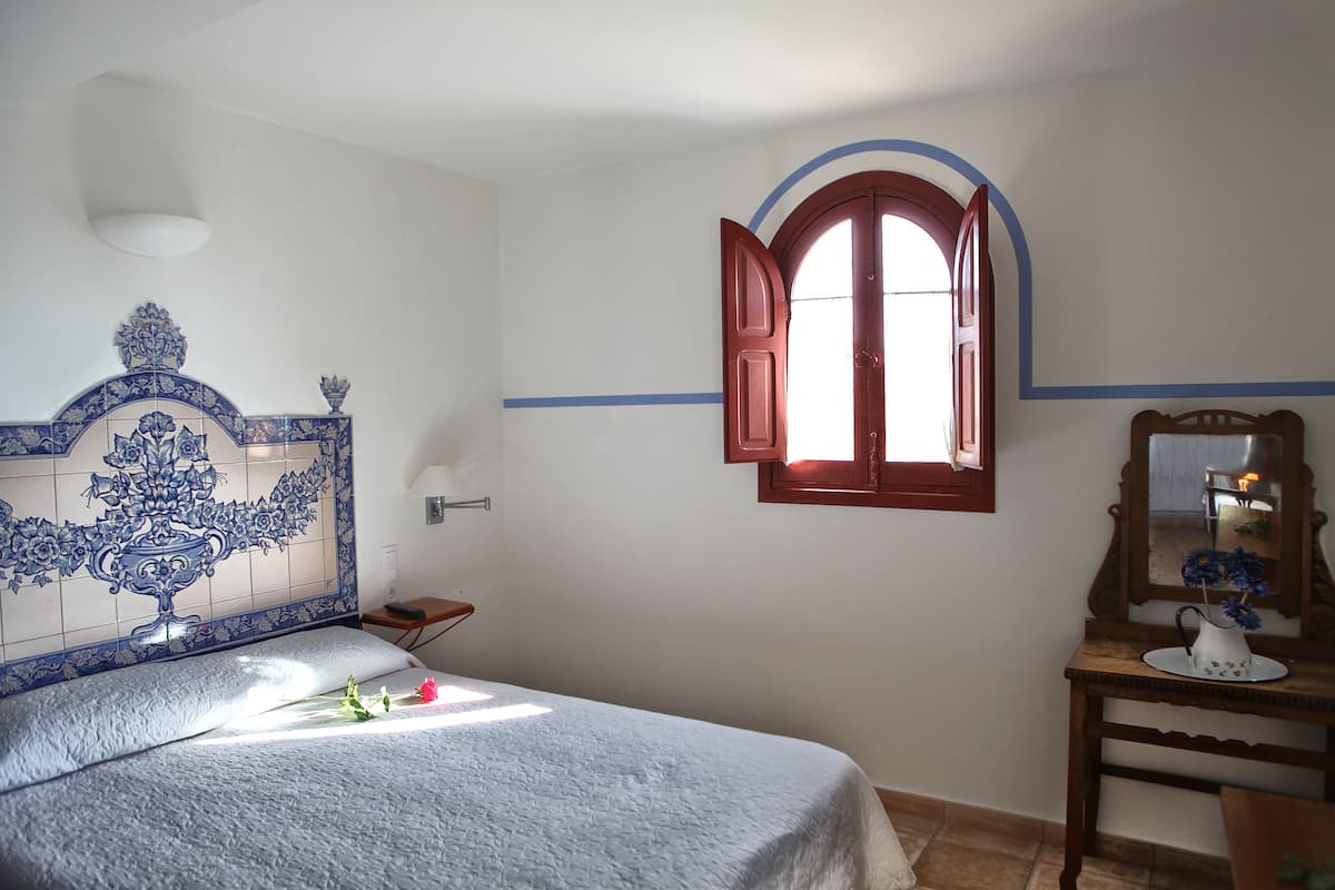 Un dormitorio con cama, mesita de noche, cómoda, espejo y ventana con contraventanas abiertas.