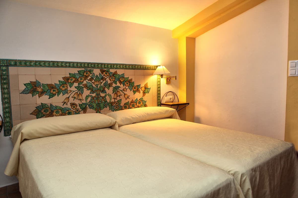 Habitación de hotel con dos camas, un adorno de pared con estampado floral, una lámpara de pared y una mesita con un reloj.