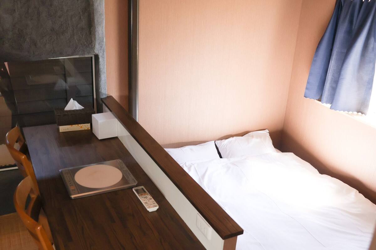 A small, compact room with a bed, a desk, and a chair. There is a window with curtains, a wall-mounted shelf, and a bedside table with a remote control.