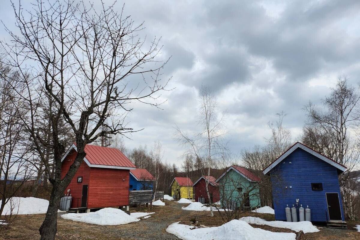 A row of small, colorful cabins with red, blue, yellow, and green roofs, surrounded by bare trees and patches of snow.