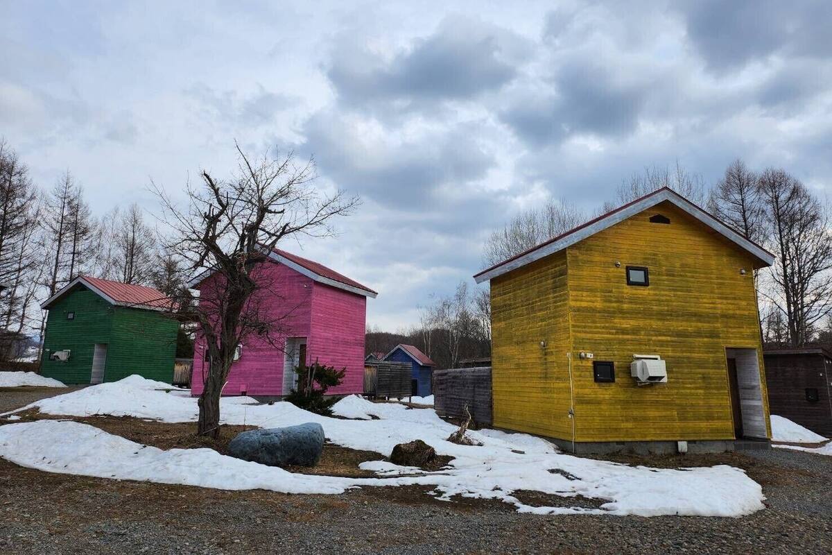 A row of colorful wooden buildings with a gravel path and snow patches in front.