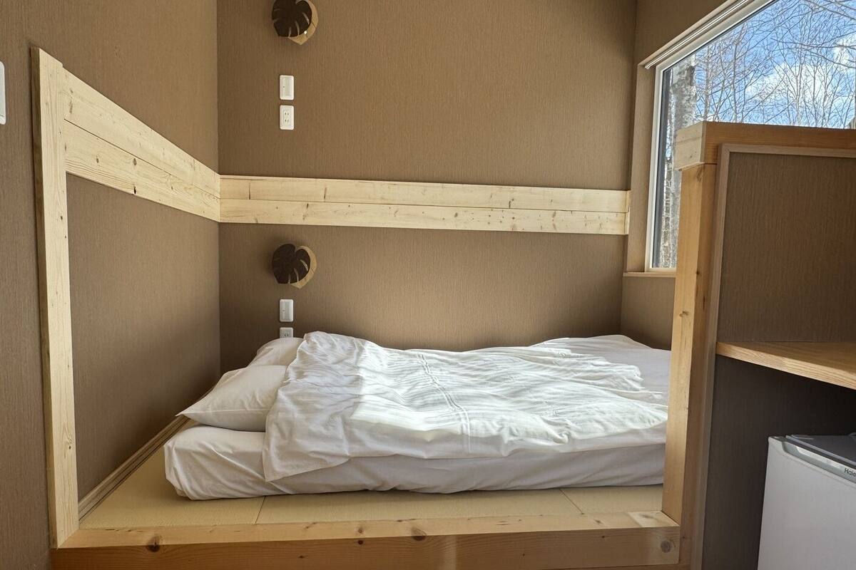 A small, single-bed room with a wooden bunk bed structure, a window with a view of trees, and a white heater on the right.