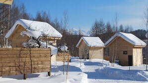 A row of wooden cabins with snow-covered roofs, surrounded by a wooden fence and trees, under a clear blue sky. - Auberge Ninguruforet (Biei)
