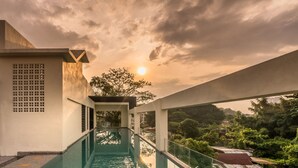 Una piscina moderna con barrera de vidrio, rodeada por un edificio blanco con diseño geométrico, y vista al atardecer sobre un paisaje verde.