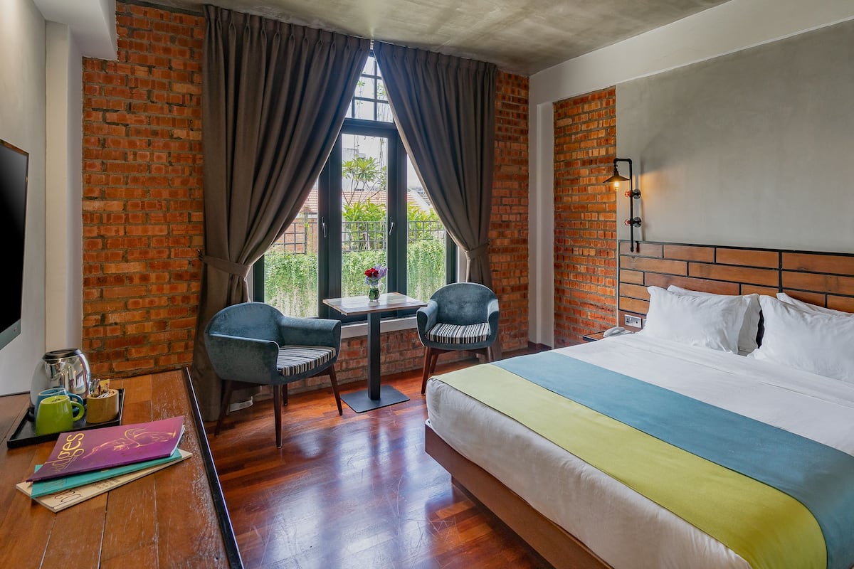 Ein Hotelzimmer mit Backsteinwand, einem Bett mit weißer und grüner Bettwäsche, einem Schreibtisch mit zwei Stühlen, einem Fenster mit Vorhängen und einem Fernseher links.