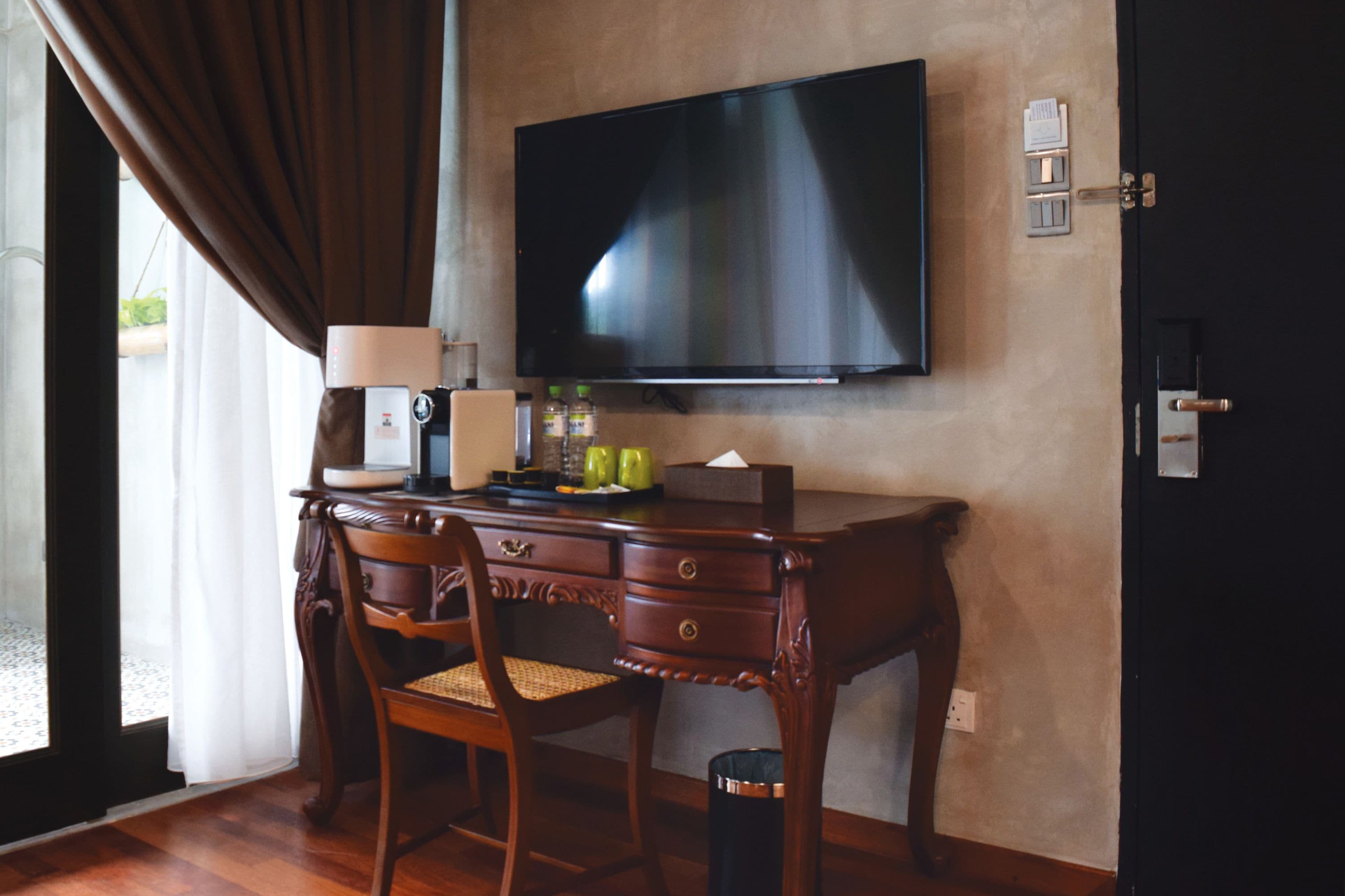 A hotel room with a wooden desk, two chairs, a flat-screen TV mounted on the wall, a lamp, and a trash can.