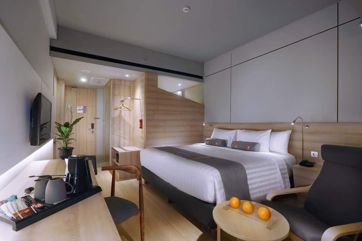 Una habitación de hotel moderna con una cama, un escritorio con cafetera, una silla, una mesita con naranjas y un televisor montado en la pared.